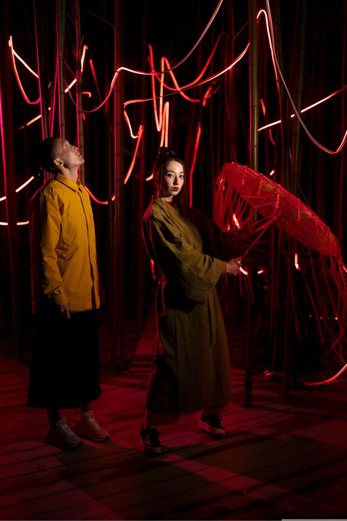 Two models in a bamboo grove lit by red neon light trails