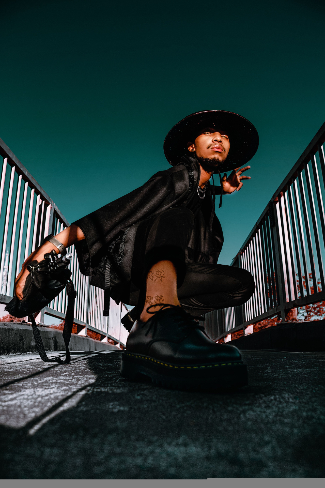 Model in all-black tailoring and wide-brim hat crouched on a Tokyo footbridge